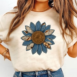 May include: A cream-colored t-shirt featuring a large, detailed sunflower design. The sunflower has blue denim petals, brown lace accents, and a brown center. The design is centered on the shirt, creating a rustic, country aesthetic.