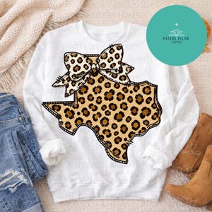 May include: A white long-sleeve sweatshirt featuring a central graphic of the state of Texas filled with a brown and black leopard print pattern, topped with a matching leopard print bow. The design has a black outline and stitch-like details.
