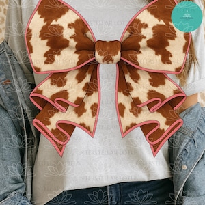 Può includere: Un grande fiocco con un motivo a pelle di mucca nei toni del marrone e crema, bordato da un bordo rosa. Il fiocco è l'elemento centrale, su uno sfondo grigio chiaro. Si tratta di un accessorio di moda.