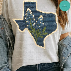 May include: A light gray t-shirt featuring a blue Texas-shaped patch with a bluebonnet flower design. The patch has a textured appearance with a cream-colored border. The shirt is paired with a denim jacket.