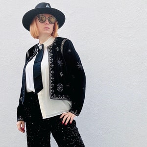 Può includere: Una donna indossa una giacca di velluto nero con ricami argentati, una blusa bianca con una cravatta nera e pantaloni neri di paillettes. Indossa un cappello nero e occhiali da sole.