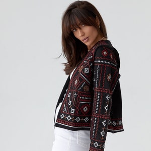 Peut inclure: Une veste noire brodée avec des motifs géométriques rouges et blancs. La veste a des manches longues et une fermeture à boutons. Le mannequin porte un pantalon blanc et un sac à main matelassé noir avec une bandoulière en chaîne dorée.