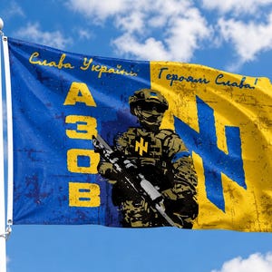 Può includere: Una bandiera blu e gialla con le parole "Slava Ukraini" e "Heroyam Slava" in scrittura gialla. La bandiera presenta una grafica di un soldato e la parola "AZOV" in lettere gialle. Lo sfondo è un cielo blu con nuvole.