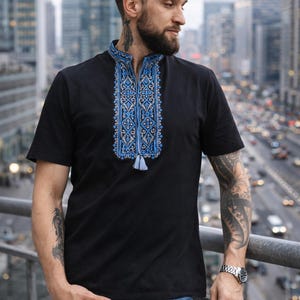 May include: A black short-sleeved shirt featuring a mandarin collar and a vertical panel of intricate blue and grey geometric embroidery down the center chest, finished with a small light blue tassel at the bottom of the embroidered design.