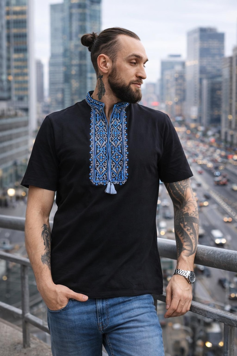 May include: A black short-sleeved shirt featuring a mandarin collar and a vertical panel of intricate blue and grey geometric embroidery down the center chest, finished with a small light blue tassel at the bottom of the embroidered design.