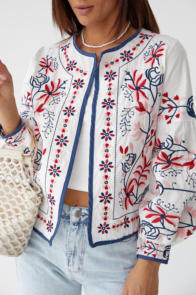 Puede incluir: Una chaqueta blanca bordada con ribete azul y patrones florales en rojo, rosa y azul.