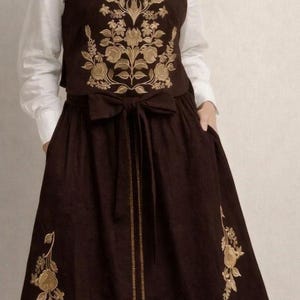 May include: A dark brown sleeveless dress featuring gold floral embroidery on the bodice and skirt, paired with a white collared shirt underneath. The dress includes a waist tie with a bow and a vertical gold line detail down the center front.