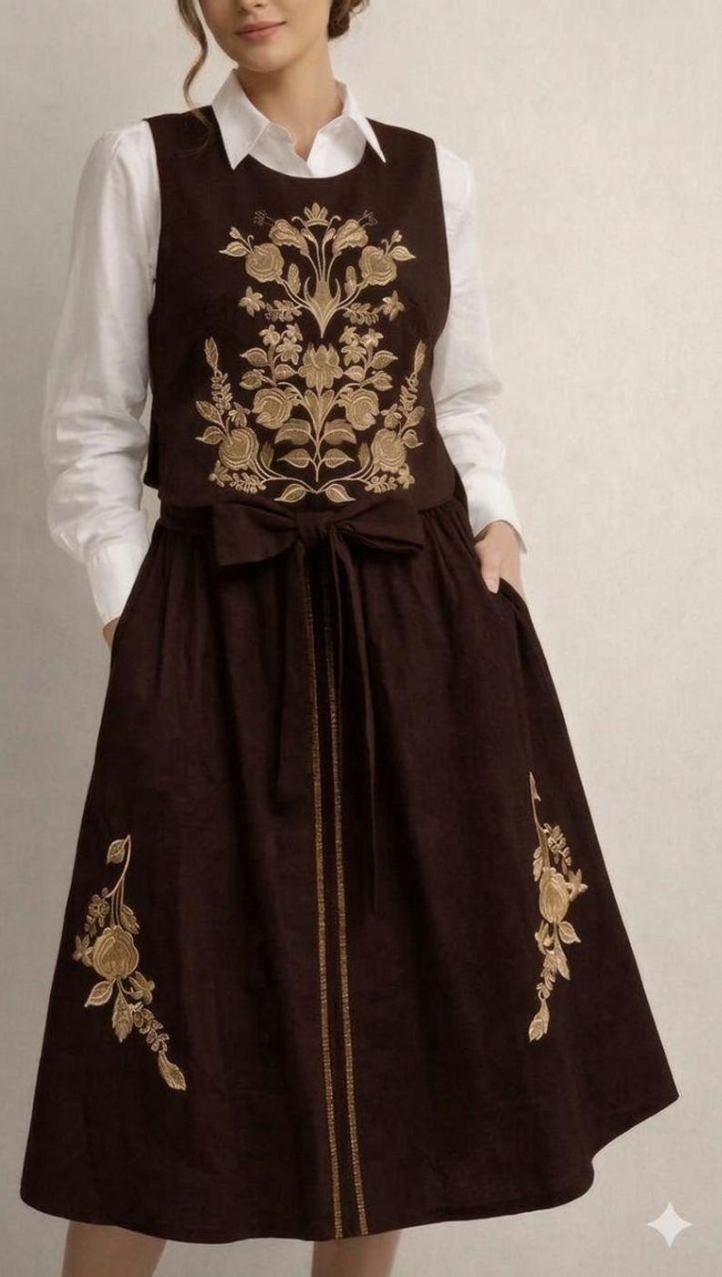 May include: A dark brown sleeveless dress featuring gold floral embroidery on the bodice and skirt, paired with a white collared shirt underneath. The dress includes a waist tie with a bow and a vertical gold line detail down the center front.
