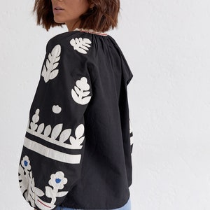 Pode incluir: Uma blusa preta com desenhos florais bordados em branco nas mangas e no decote. A blusa tem um caimento solto e um decote ligeiramente franzido.
