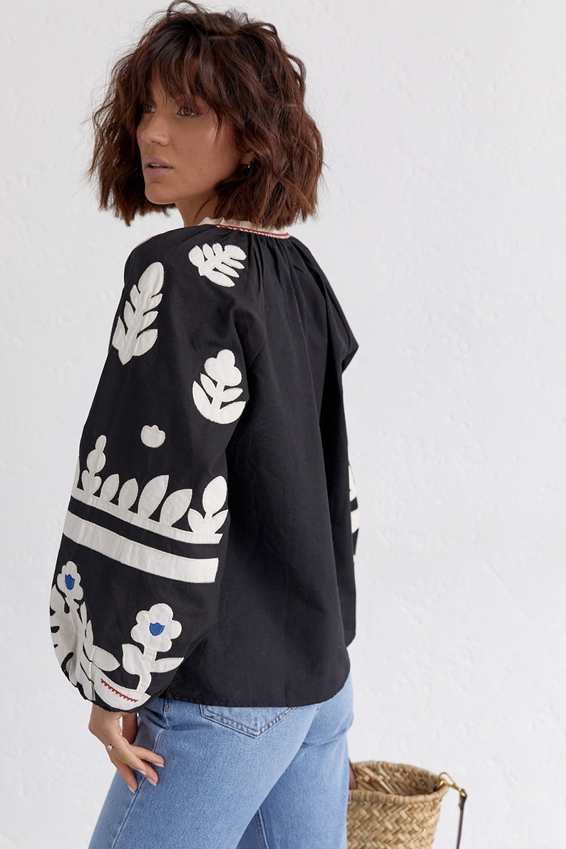Pode incluir: Uma blusa preta com desenhos florais bordados em branco nas mangas e no decote. A blusa tem um caimento solto e um decote ligeiramente franzido.