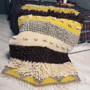 Può includere: Una coperta tessuta multicolore con un bordo a frangia. La coperta presenta strisce di colore giallo, marrone, bianco e grigio. La coperta è realizzata in un materiale morbido e soffice.