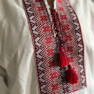 May include: A white linen shirt with intricate red and white embroidery around the collar and chest. The embroidery features geometric patterns and is accented by red tassels. The shirt has a mandarin collar and a relaxed fit.