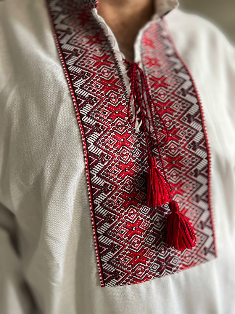 May include: A white linen shirt with intricate red and white embroidery around the collar and chest. The embroidery features geometric patterns and is accented by red tassels. The shirt has a mandarin collar and a relaxed fit.