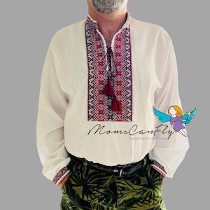 May include: White long-sleeve shirt with intricate red and black embroidery on the collar and chest. The shirt features a tassel tie and embroidered cuffs. The text "MomsCanFly" is printed on the shirt.