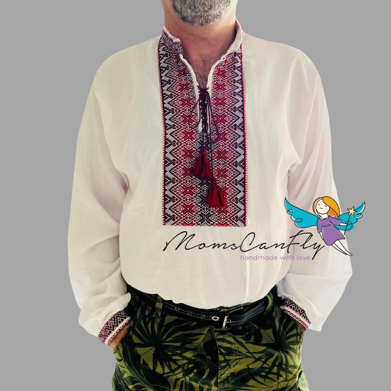 May include: White long-sleeve shirt with intricate red and black embroidery on the collar and chest. The shirt features a tassel tie and embroidered cuffs. The text "MomsCanFly" is printed on the shirt.