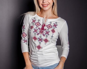 May include: A woman wearing a white long-sleeved shirt with a floral embroidered design and blue jeans. She is sitting on a white stool.