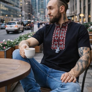 May include: A black short-sleeved shirt featuring a stand-up collar and a central vertical panel with intricate red, white, and black geometric embroidery. The neckline includes a small slit with two hanging gray tassels at the bottom of the design.