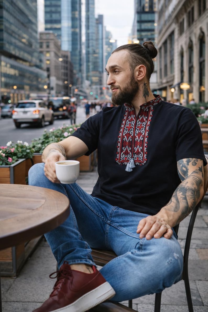 May include: A black short-sleeved shirt featuring a stand-up collar and a central vertical panel with intricate red, white, and black geometric embroidery. The neckline includes a small slit with two hanging gray tassels at the bottom of the design.