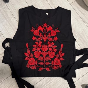 May include: A black sleeveless top featuring a vibrant red floral embroidery pattern centered on the front. The garment has a round neckline and long black fabric ties extending from both sides of the waist for an adjustable fit.