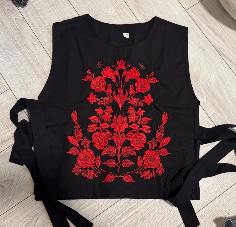 May include: A black sleeveless top featuring a vibrant red floral embroidery pattern centered on the front. The garment has a round neckline and long black fabric ties extending from both sides of the waist for an adjustable fit.
