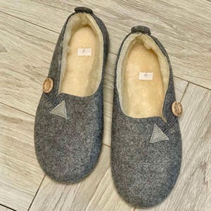 May include: A pair of gray felt slippers with a light-colored interior. Each slipper has a wooden button and a small gray triangle detail. The slippers are size 40.