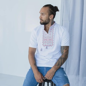 May include: A white short-sleeved shirt featuring a vertical panel of intricate red and blue embroidery along the center neckline. The shirt includes a split collar design with two small hanging tassels at the base of the embroidered pattern.