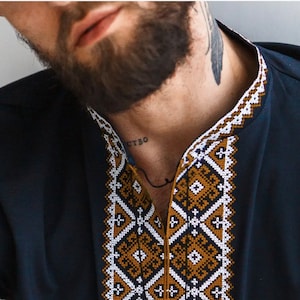 May include: Navy blue shirt with intricate geometric embroidery in white, gold, and dark blue along the collar and neckline. The shirt has a classic, tailored fit and a simple, elegant design.