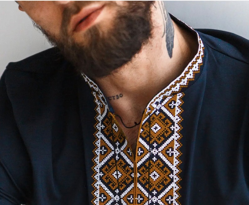May include: Navy blue shirt with intricate geometric embroidery in white, gold, and dark blue along the collar and neckline. The shirt has a classic, tailored fit and a simple, elegant design.