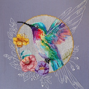 May include: A cross-stitch design of a colourful hummingbird, set within a gold-edged circle. The bird is surrounded by pink, yellow, and purple flowers, with white leaf accents, against a light purple background. The design showcases detailed needlework.