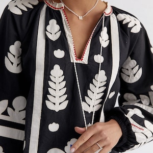 Pode incluir: Uma blusa preta de manga comprida com desenhos florais bordados em branco. A blusa tem um decote em V e uma borda vermelha ao redor do decote. A modelo est&aacute; usando um colar de prata com um pequeno pingente em forma de letra.