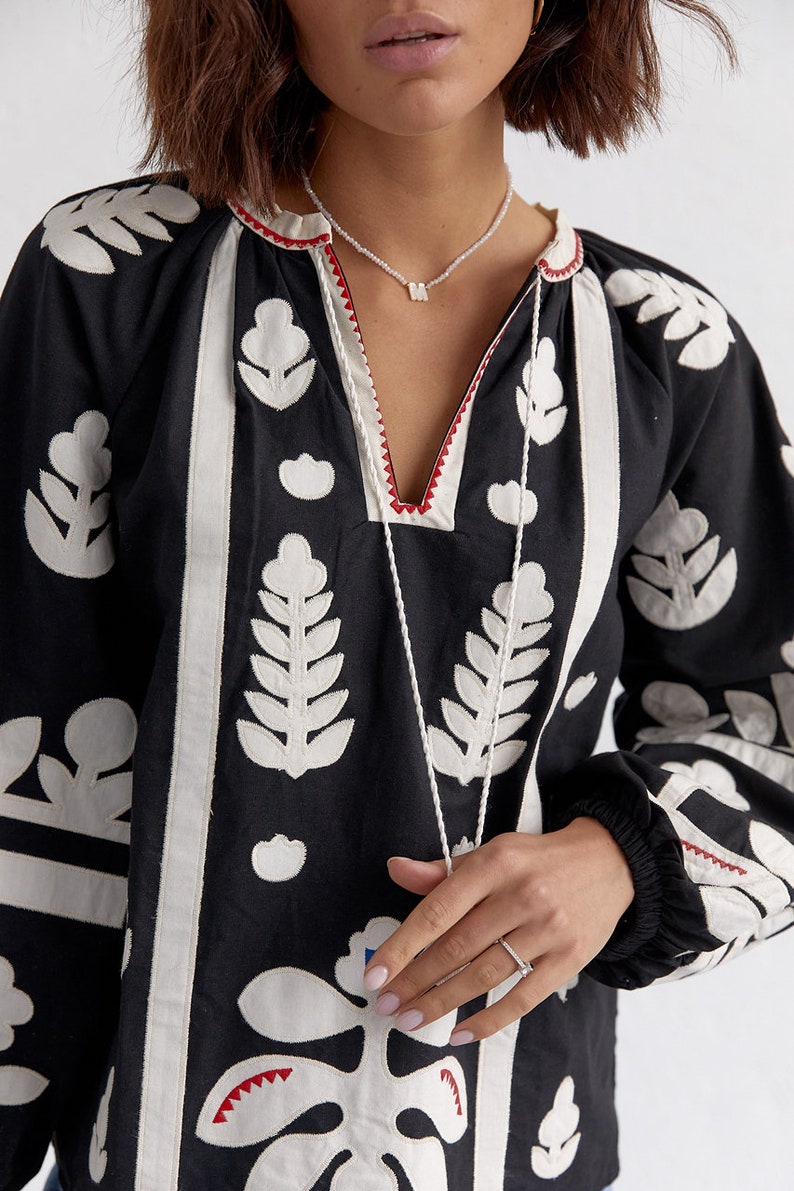 Pode incluir: Uma blusa preta de manga comprida com desenhos florais bordados em branco. A blusa tem um decote em V e uma borda vermelha ao redor do decote. A modelo est&aacute; usando um colar de prata com um pequeno pingente em forma de letra.