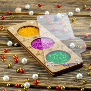 Può includere: Organizzatore di perline in legno con tre scomparti riempiti di perline gialle, viola e verdi. Un coperchio in acrilico trasparente è parzialmente aperto, rivelando altre perline. L'organizzatore è decorato con disegni di libellule incisi.