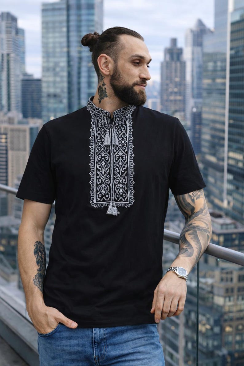 May include: A black short-sleeved shirt featuring a mandarin collar and a central vertical panel with intricate white geometric embroidery. The neckline includes a small slit with two hanging grey tassels, creating a detailed contrast against the fabric.