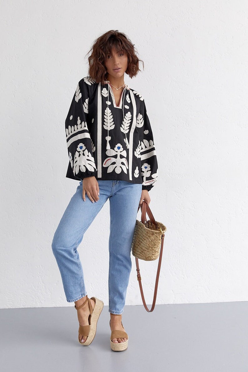 Pode incluir: Uma blusa bordada preta e branca com um padr&atilde;o floral. A blusa tem mangas compridas e um decote em V. A modelo est&aacute; usando jeans azul e sand&aacute;lias de plataforma marrons.