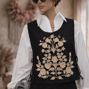 May include: A black sleeveless vest featuring intricate beige floral embroidery on the front, with side lace-up closures, worn over a crisp white long-sleeved collared shirt, paired with a matching black skirt and a small grey handbag.