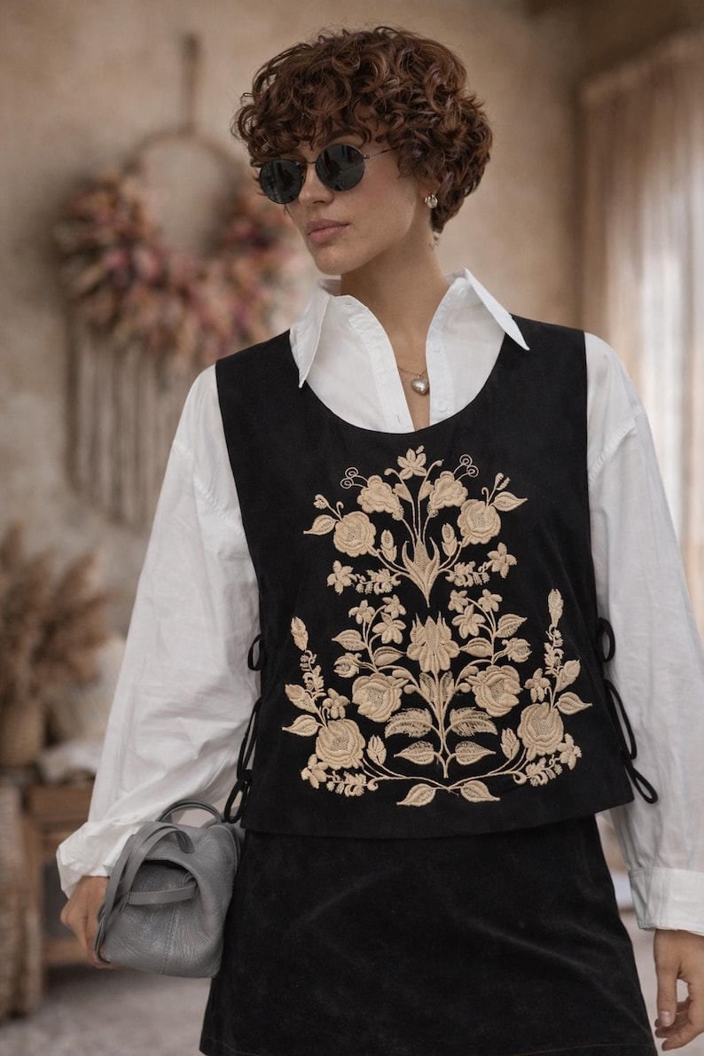 May include: A black sleeveless vest featuring intricate beige floral embroidery on the front, with side lace-up closures, worn over a crisp white long-sleeved collared shirt, paired with a matching black skirt and a small grey handbag.