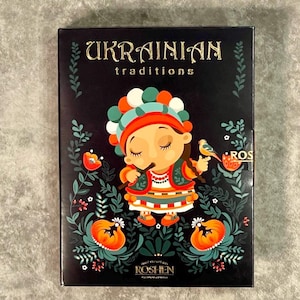 May include: A black box with the words "UKRAINIAN traditions" in gold lettering. The box features a cartoon illustration of a person in traditional Ukrainian clothing, holding a flute and a bird. The box is decorated with floral and leaf motifs.