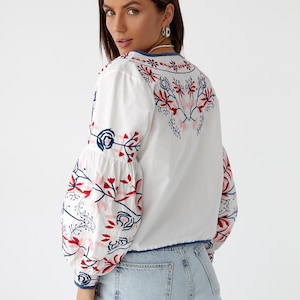 Puede incluir: Blusa blanca de manga larga con un dise&ntilde;o de bordado floral en rojo, azul y rosa. La blusa tiene cuello en V y un corte ligeramente recortado.
