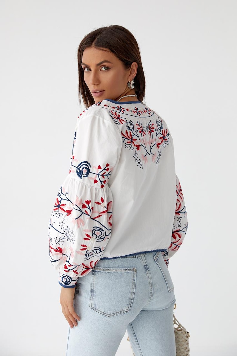 Puede incluir: Blusa blanca de manga larga con un dise&ntilde;o de bordado floral en rojo, azul y rosa. La blusa tiene cuello en V y un corte ligeramente recortado.