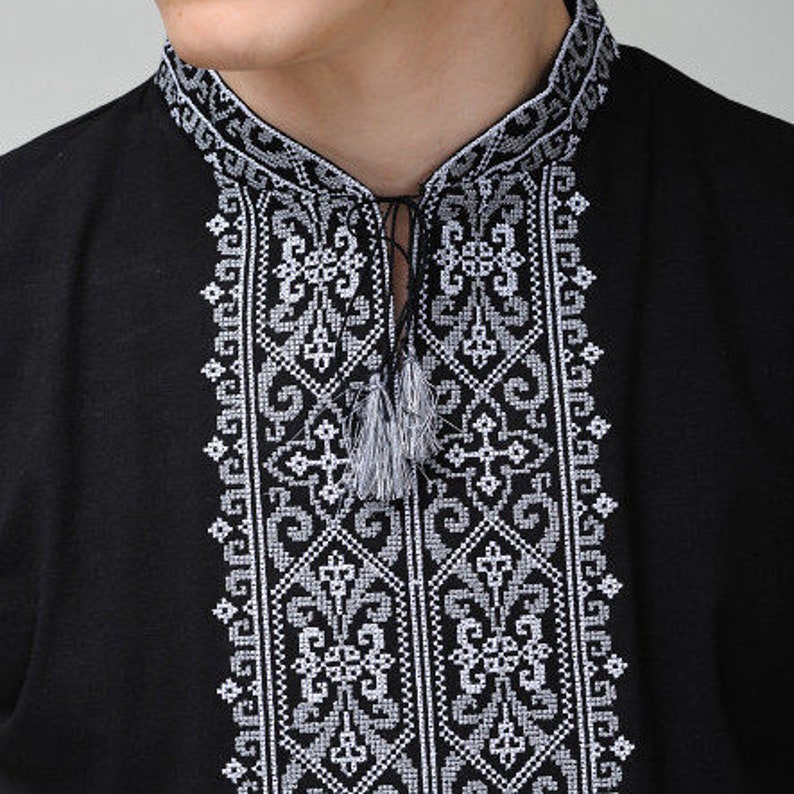 May include: A black shirt featuring a mandarin collar and a vertical panel of intricate white geometric embroidery down the center. The neckline includes a thin black cord with two small, light gray tassels hanging from the front.