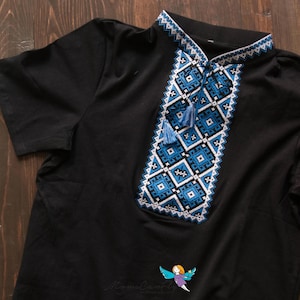 May include: Black short-sleeved shirt with intricate blue and white embroidered geometric patterns on the collar and chest. The design includes tassels and a small logo at the bottom. The shirt is made of a soft material.