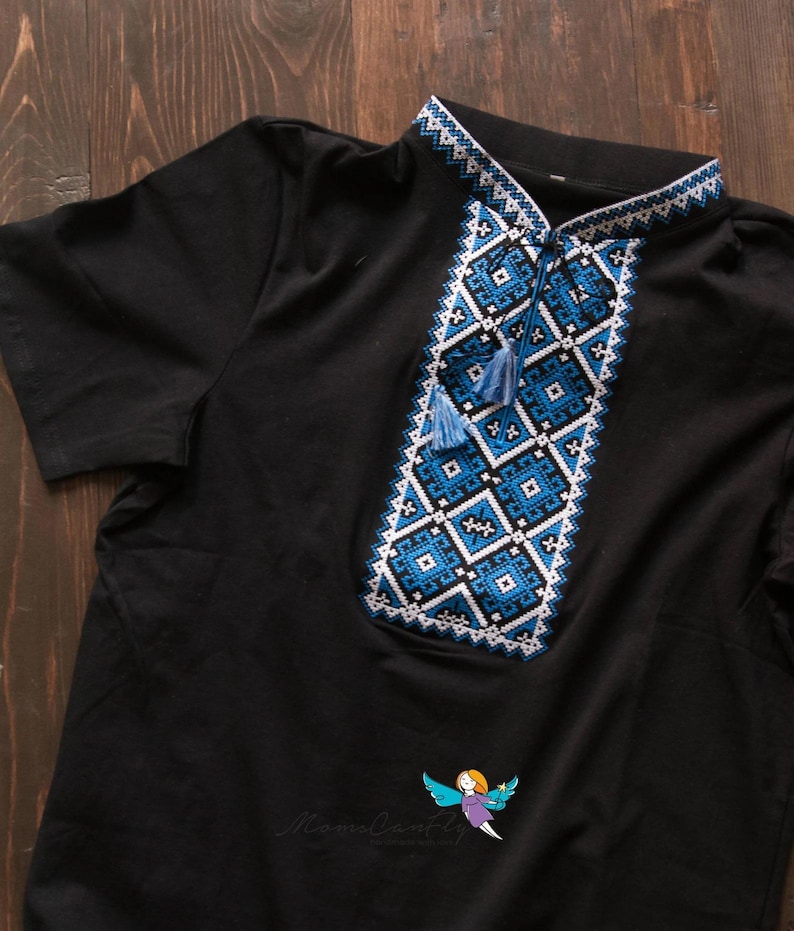 May include: Black short-sleeved shirt with intricate blue and white embroidered geometric patterns on the collar and chest. The design includes tassels and a small logo at the bottom. The shirt is made of a soft material.