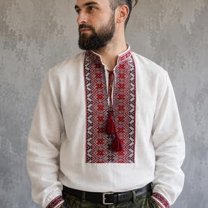 May include: White linen shirt with long sleeves, featuring intricate red and black geometric embroidery on the chest and cuffs. The shirt has a mandarin collar and a tassel detail. The model is wearing green camouflage pants.