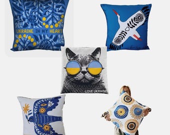 Puede incluir: Cinco cojines decorativos con varios diseños. Uno presenta un estampado floral azul con el texto "UKRAINE IN MY HEART". Otro muestra una cigüeña blanca sobre fondo azul. Un tercer cojín muestra un gato con gafas de sol con el texto "LOVE UKRAINE."