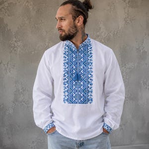 Puede incluir: Camisa blanca de manga larga con intrincados bordados azules en la parte delantera y en los puños. El bordado presenta patrones geométricos y una borla en el escote. La camisa se combina con vaqueros de mezclilla de lavado claro.