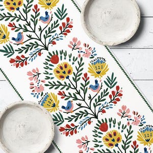 May include: White table runner with a floral design featuring yellow, red, and pink flowers, green leaves, and blue birds. The runner has a green stitched border and is paired with two white ceramic plates.