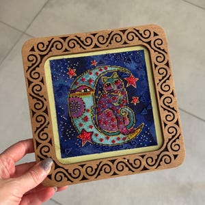 Peut inclure: Une broderie au point de croix représentant un chat et une lune sur un fond bleu étoilé. L'œuvre est encadrée dans un cadre en bois décoratif avec un motif de volutes. Le chat est violet et rouge, et la lune est bleu clair. Le cadre mesure environ 15 cm.