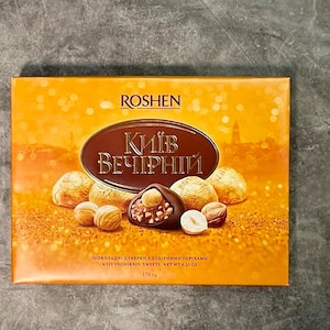 May include: A box of Kyiv Vechirniy sweets by Roshen. The box is orange with a brown oval in the center with text. Several candies are visible, some with hazelnuts. The net weight is 6.21 oz.