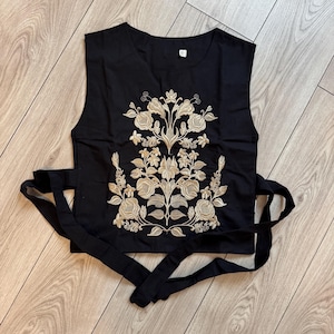 May include: A black sleeveless top featuring a large, intricate gold floral embroidery pattern centered on the front. The garment includes a round neckline and long black fabric ties extending from the sides for a wrap-around closure.