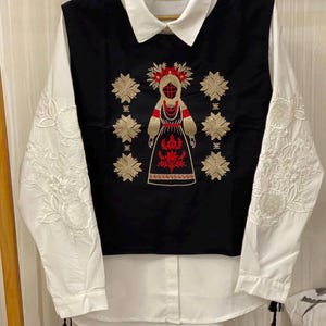 May include: A long-sleeved white shirt featuring a black vest overlay. The black section displays intricate gold and red embroidery of a central figure surrounded by geometric star patterns. The white sleeves are adorned with detailed white floral embroidery.